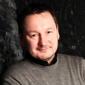 Ein Mann mit kurzen dunklen Haaren und hellem Bartstoppeln, gekleidet in einen grauen Pullover über einem schwarzen Hemd, lächelt sanft, während er vor einem dunklen, strukturierten Hintergrund steht und den zugänglichen Stil eines professionellen Contact Center-Beraters verkörpert. - RUF Beratung - Ihre Contact Center Berater