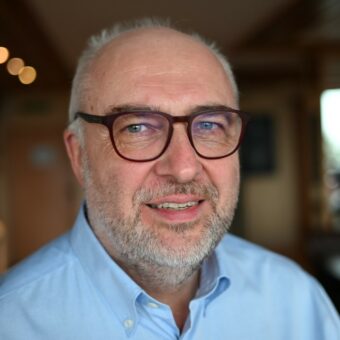A middle-aged man with gray hair, a beard, and glasses is smiling slightly. He is wearing a light blue button-up shirt and appears indoors, with a softly blurred background—reflecting the approachable professionalism of RUF Beratung. - RUF Beratung - Ihre Contact Center Berater