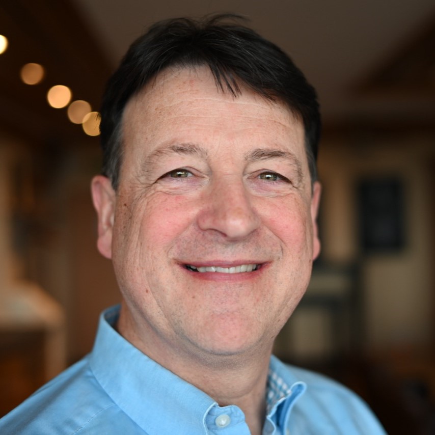 Thomas Ulrich mit kurzen dunklen Haaren lächelt warm in die Kamera. Sein hellblaues Hemd mit Kragen strahlt Professionalität aus – perfekt für einen Contact Center Berater –, während der sanft verschwommene, warm beleuchtete Hintergrund eine einladende Note verleiht. - RUF Beratung - Ihre Contact Center Berater