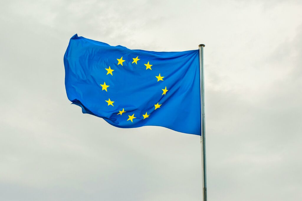 Eine blaue Flagge mit einem Kreis aus zwölf gelben Sternen, die die Europäische Union repräsentieren, weht an einem Fahnenmast vor einem bewölkten Himmel – ein ideales Bild für den Hintergrund eines Kundenservice-Workshops zum EU AI Act. - RUF Beratung - Ihre Contact Center Berater