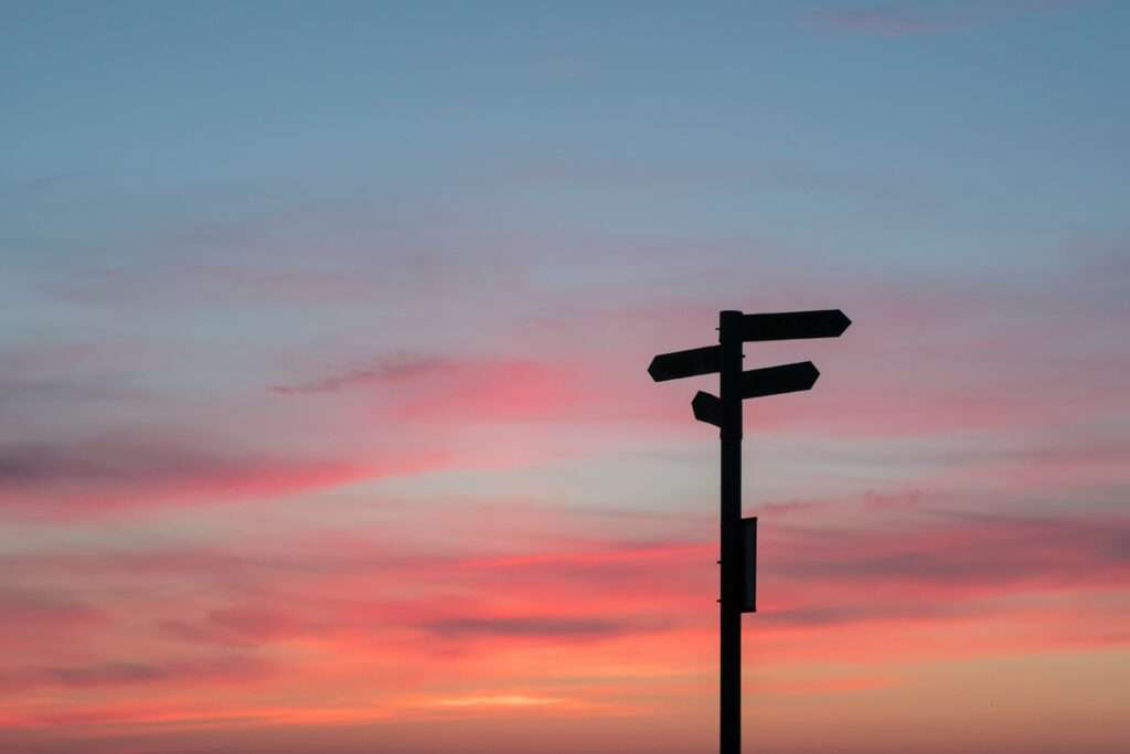 Vor einem farbenfrohen Sonnenuntergangshimmel steht die Silhouette eines Wegweisers mit mehreren Richtungspfeilen und symbolisiert inmitten sanfter Rosa-, Orange- und Blautöne das Änderungsmanagement. - RUF Beratung - Ihre Contact Center Berater