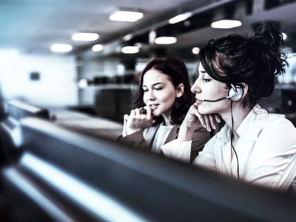 Zwei Frauen in einem Büro, eine davon mit Headset, sitzen nebeneinander vor Computerbildschirmen und wirken konzentriert und engagiert in ihrer Kundenservice-Arbeit. Der Hintergrund ist unscharf. - RUF Beratung - Ihre Contact Center Berater