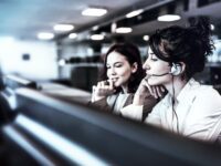 Zwei Frauen in einem Büro, eine davon mit Headset, sitzen nebeneinander vor Computerbildschirmen und wirken konzentriert und engagiert in ihrer Kundenservice-Arbeit. Der Hintergrund ist unscharf. - RUF Beratung - Ihre Contact Center Berater